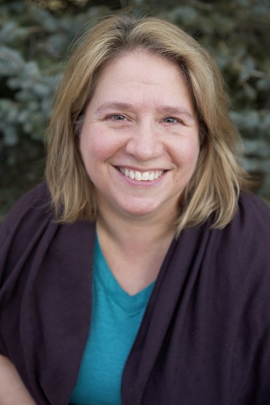 An image of Kelly, smiling, wearing a purple jacket and a light blue shirt.